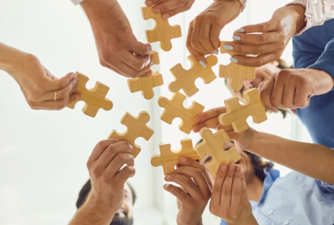 Ten hands in a circle holding a wooden jigsaw piece each in the middle